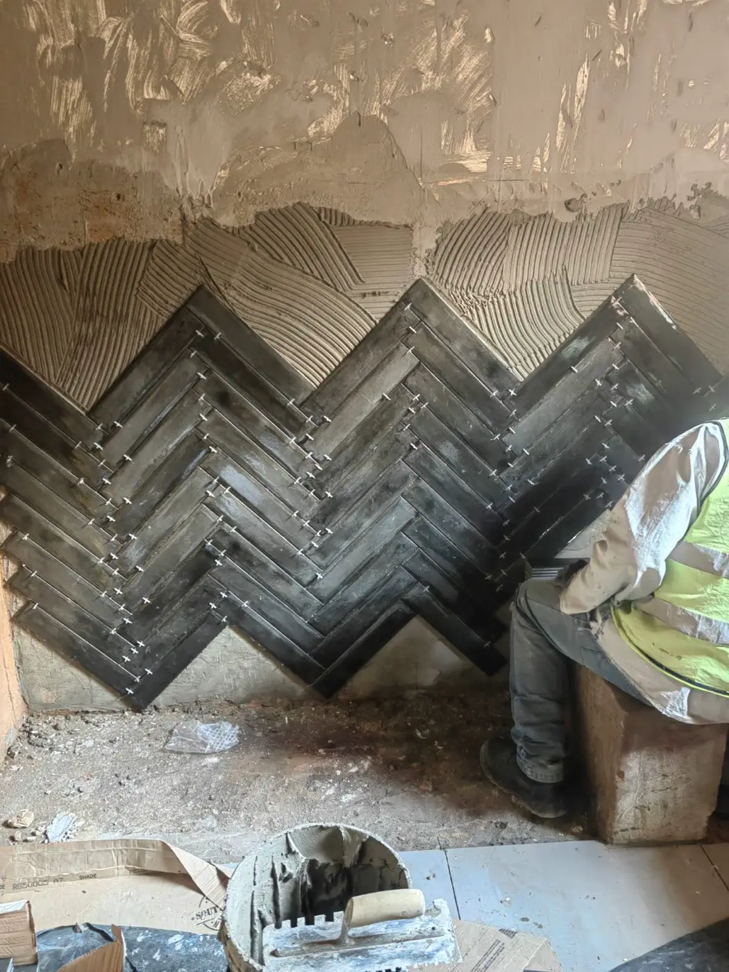 A tiler setting black herringbone tiles on a feature wall